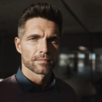 Close-up portrait of a man with dark hair and stubble, in a dim indoor setting, looking at the camera.