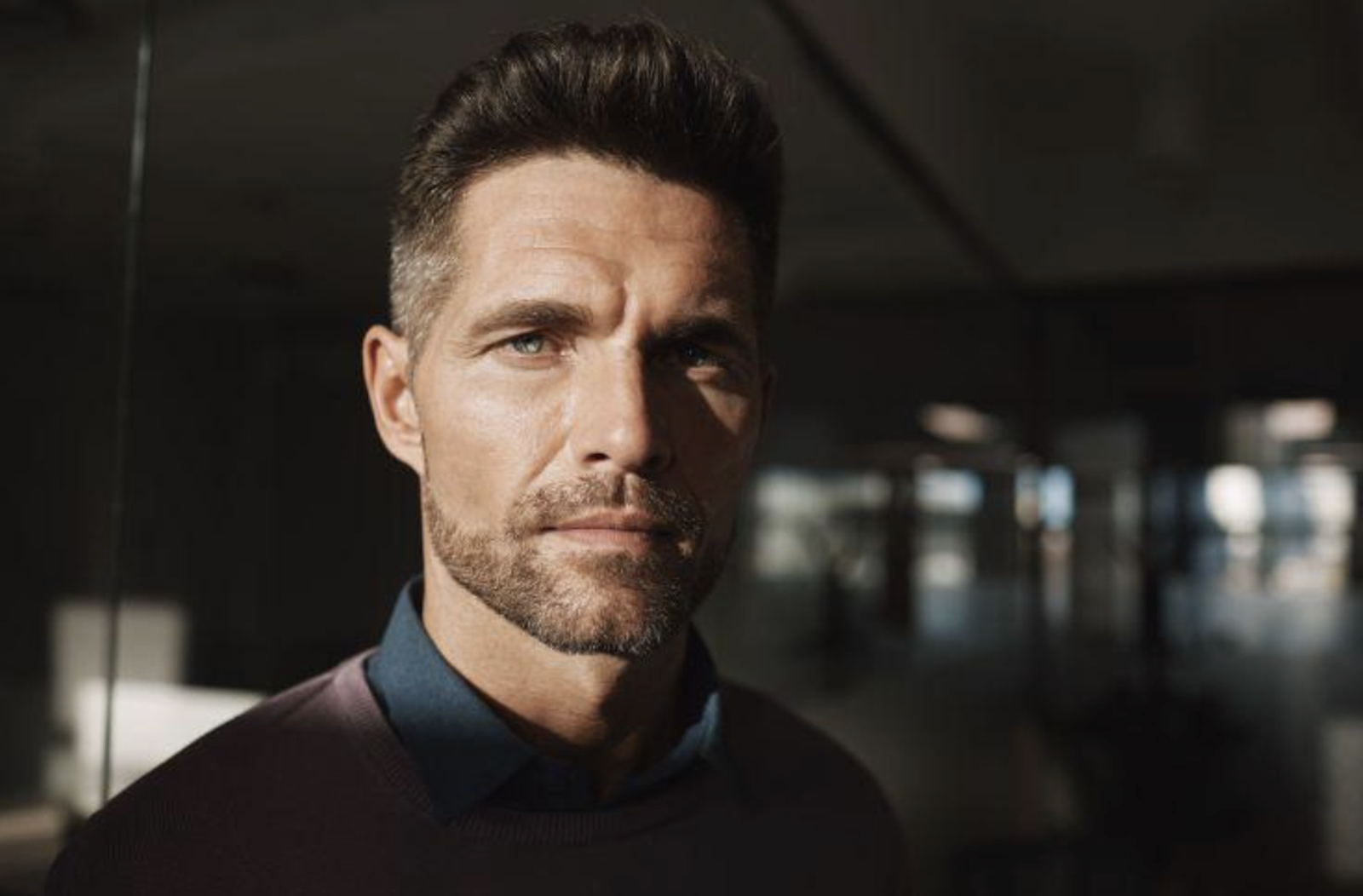 Close-up portrait of a man with dark hair and stubble, in a dim indoor setting, looking at the camera.