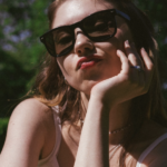 Young woman in oversized sunglasses rests her chin on her hand, lips pursed, outdoors in a sunny park.