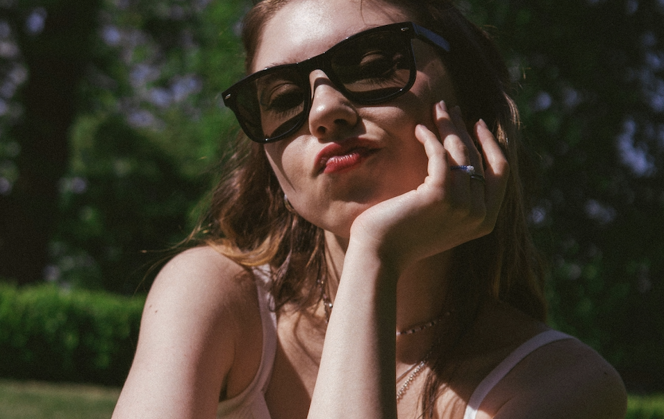 Young woman in oversized sunglasses rests her chin on her hand, lips pursed, outdoors in a sunny park.