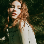 Woman with long red hair poses outdoors among trees, looking slightly toward the camera with a calm expression in sunlight.
