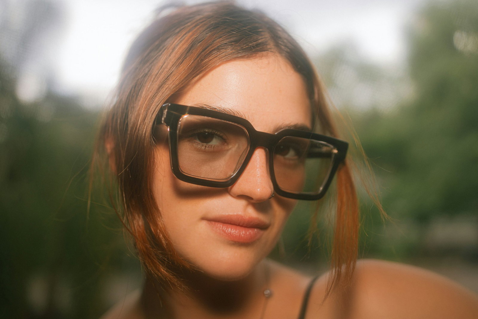 Close-up portrait of a woman with brown hair wearing oversized black glasses, outdoors with a blurred green background.