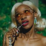 Portrait of a stylish woman with blonde hair wearing orange-tinted sunglasses, a mint green knotted strapless top, and pearl necklace outdoors at night.