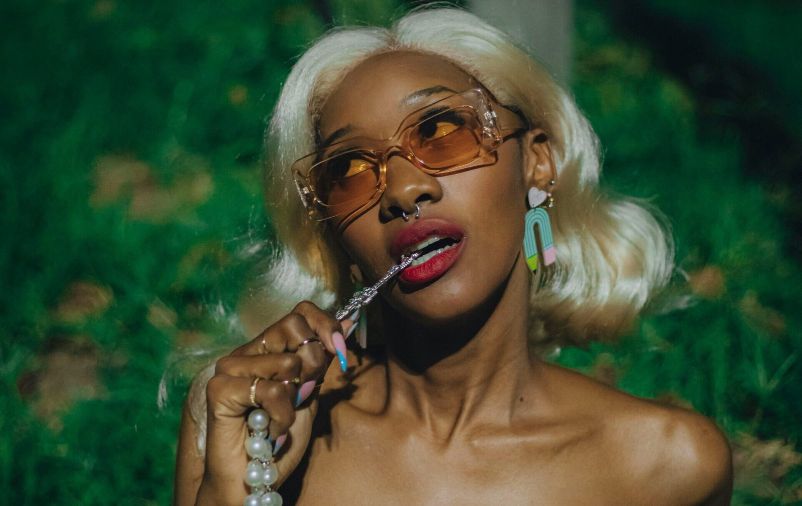Portrait of a stylish woman with blonde hair wearing orange-tinted sunglasses, a mint green knotted strapless top, and pearl necklace outdoors at night.