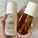 Two Phlur fragrance bottles held in a hand: white 'Heavy Cream' and amber 'Vanilla Skin' with a light fabric backdrop.