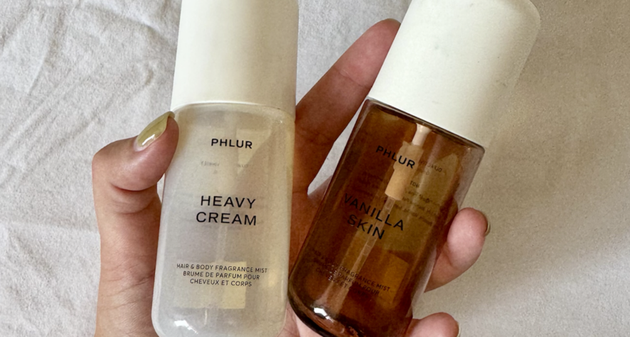 Two Phlur fragrance bottles held in a hand: white 'Heavy Cream' and amber 'Vanilla Skin' with a light fabric backdrop.