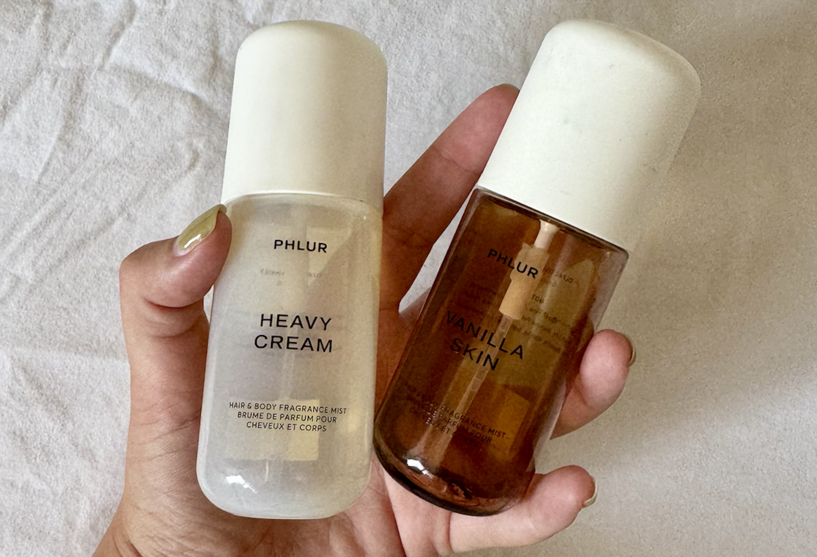 Two Phlur fragrance bottles held in a hand: white 'Heavy Cream' and amber 'Vanilla Skin' with a light fabric backdrop.