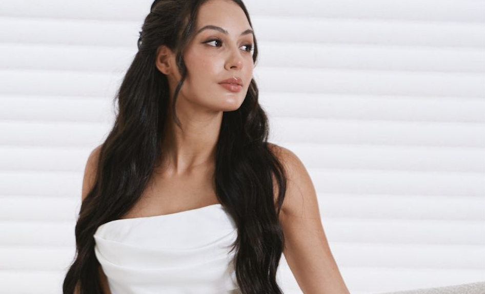 Portrait of a woman with long dark hair wearing a white strapless top, looking to the right against a light background.