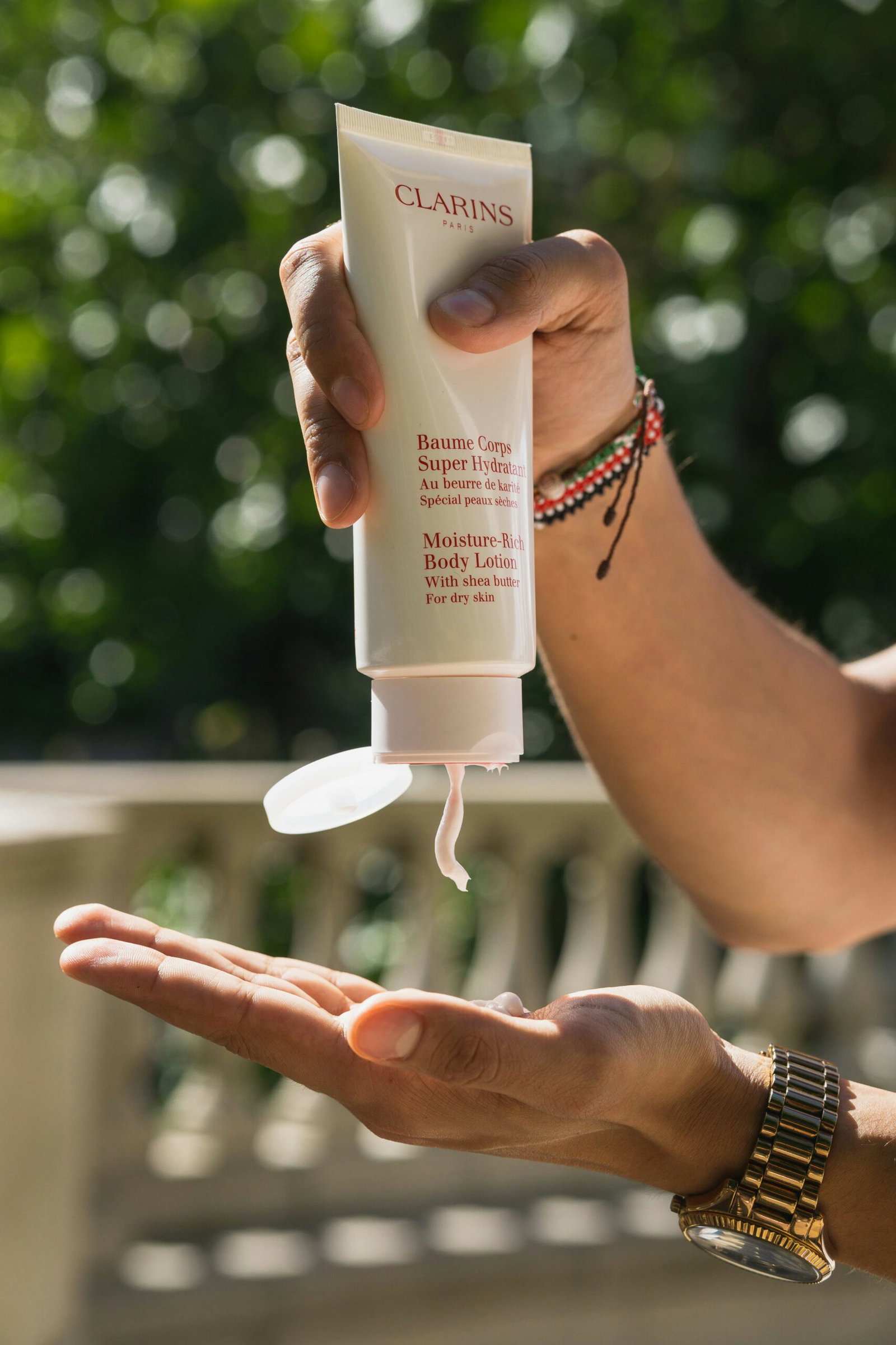 Hand squeezing Clarins Moisture-Rich Body Lotion onto an open palm outdoors, product label visible on the tube.