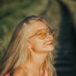 Woman with blonde hair and orange sunglasses smiling outdoors on a sunny day.