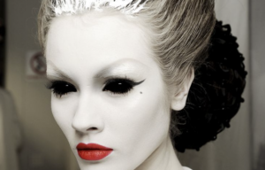 Dramatic Halloween makeup look featuring ghostly white face makeup, blacked-out eyes with winged eyeliner, long lashes, bright red lips and a beauty spot.