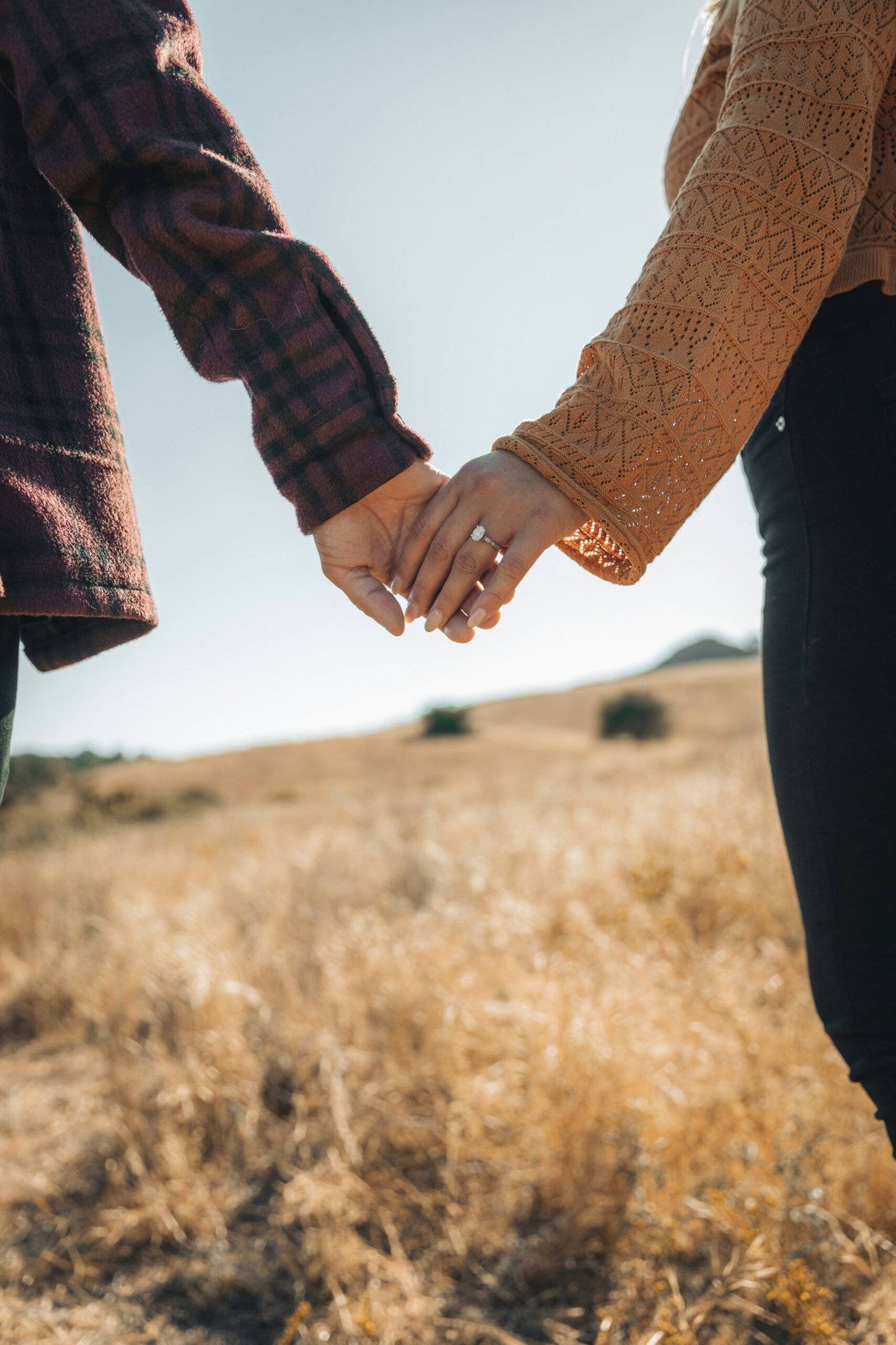 Couple holding hands outdoors, showing a diamond engagement ring and a natural nude manicure.