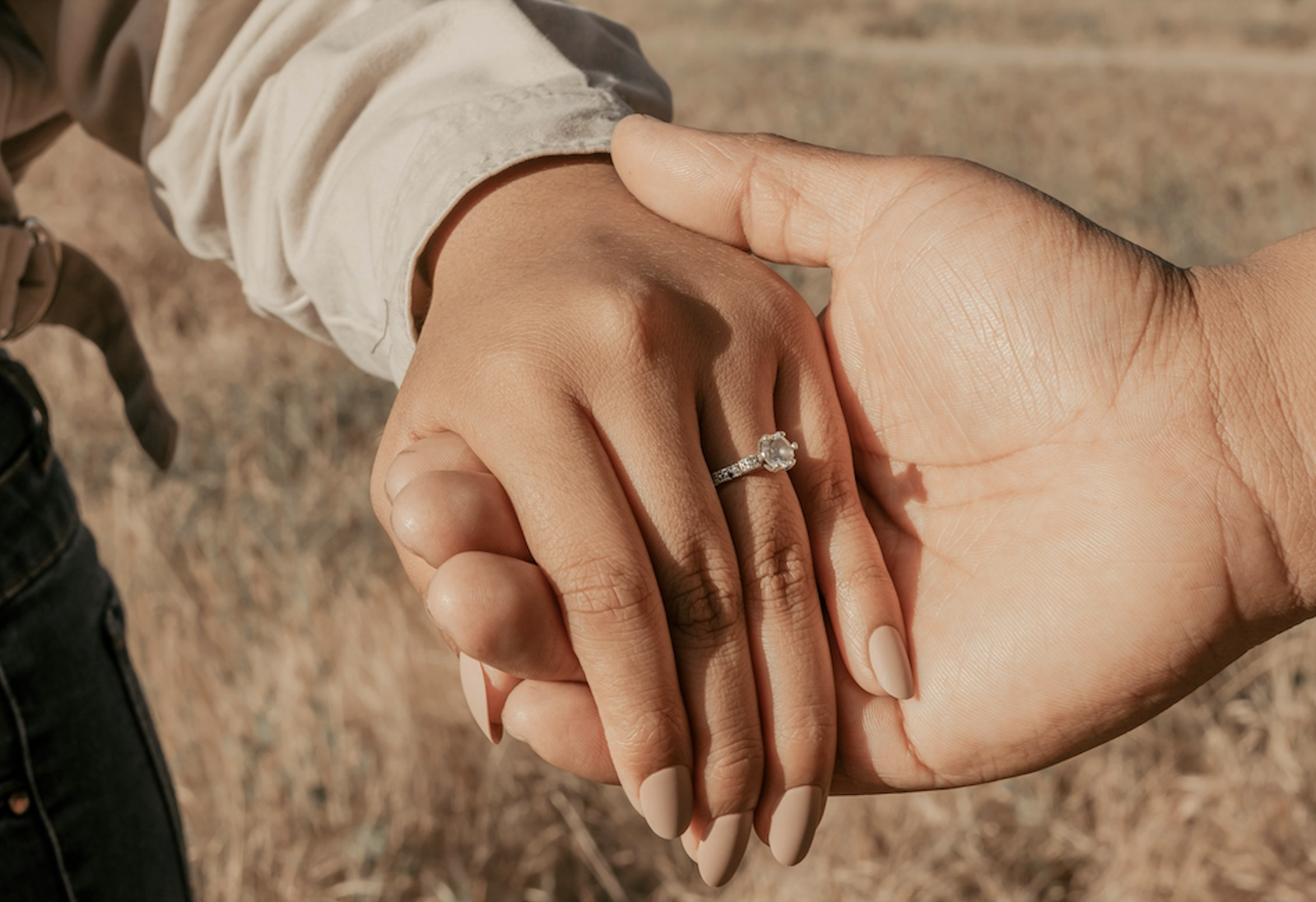 Close-up of a diamond engagement ring paired with nude almond-shaped nails as a couple holds hands.
