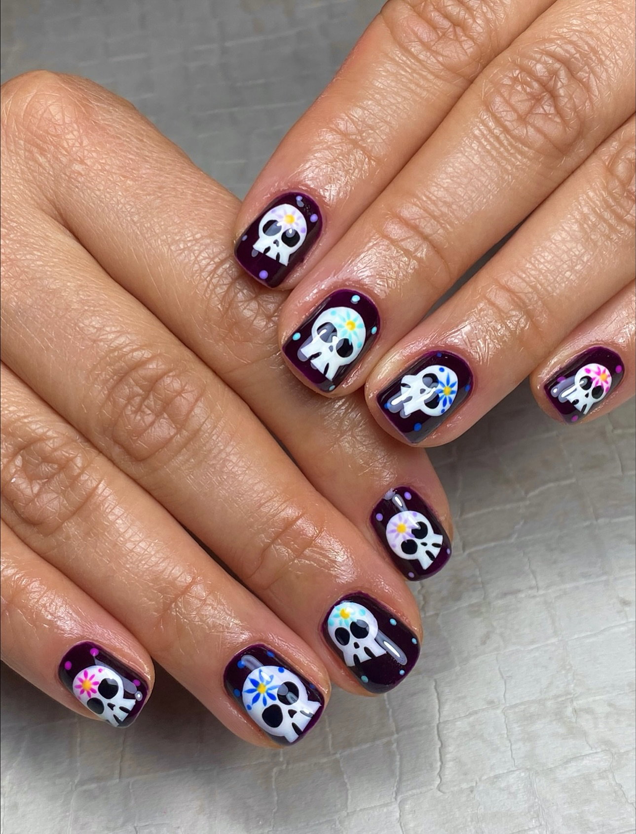 Short dark purple Halloween nails decorated with white Day of the Dead skulls and colourful flowers