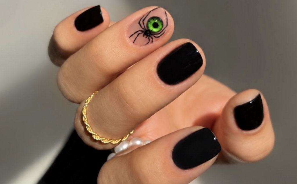 Short glossy black Halloween nails with a striking green-eyed spider design on a nude accent nail.