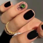Short glossy black Halloween nails with a striking green-eyed spider design on a nude accent nail.