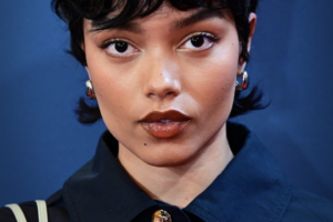 Whitney Peak in a close-up beauty portrait, wearing glossy brown lipstick and short dark hair against a deep blue background.