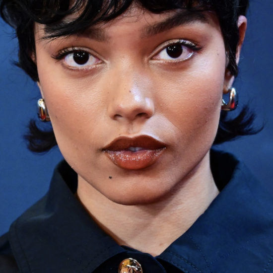 Whitney Peak in a close-up beauty portrait, wearing glossy brown lipstick and short dark hair against a deep blue background.