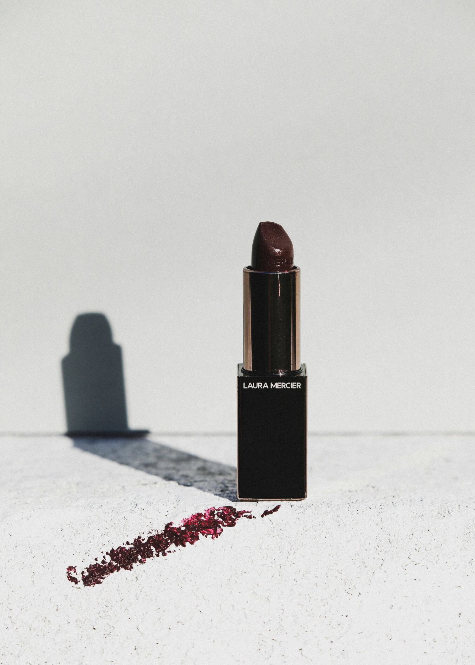 Open Laura Mercier deep berry lipstick beside a berry-coloured swatch, casting a shadow on a pale surface.