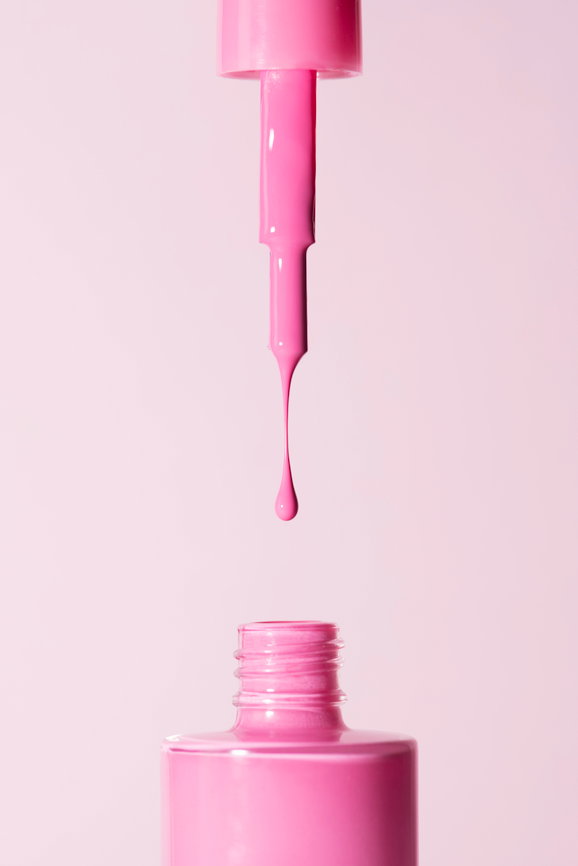 Close-up of a brush dripping bright pink nail polish above an open bottle against a pale pink background.