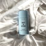 Salt & Stone Lily & Yuzu natural deodorant in pale blue packaging resting on sunlit, textured white fabric.