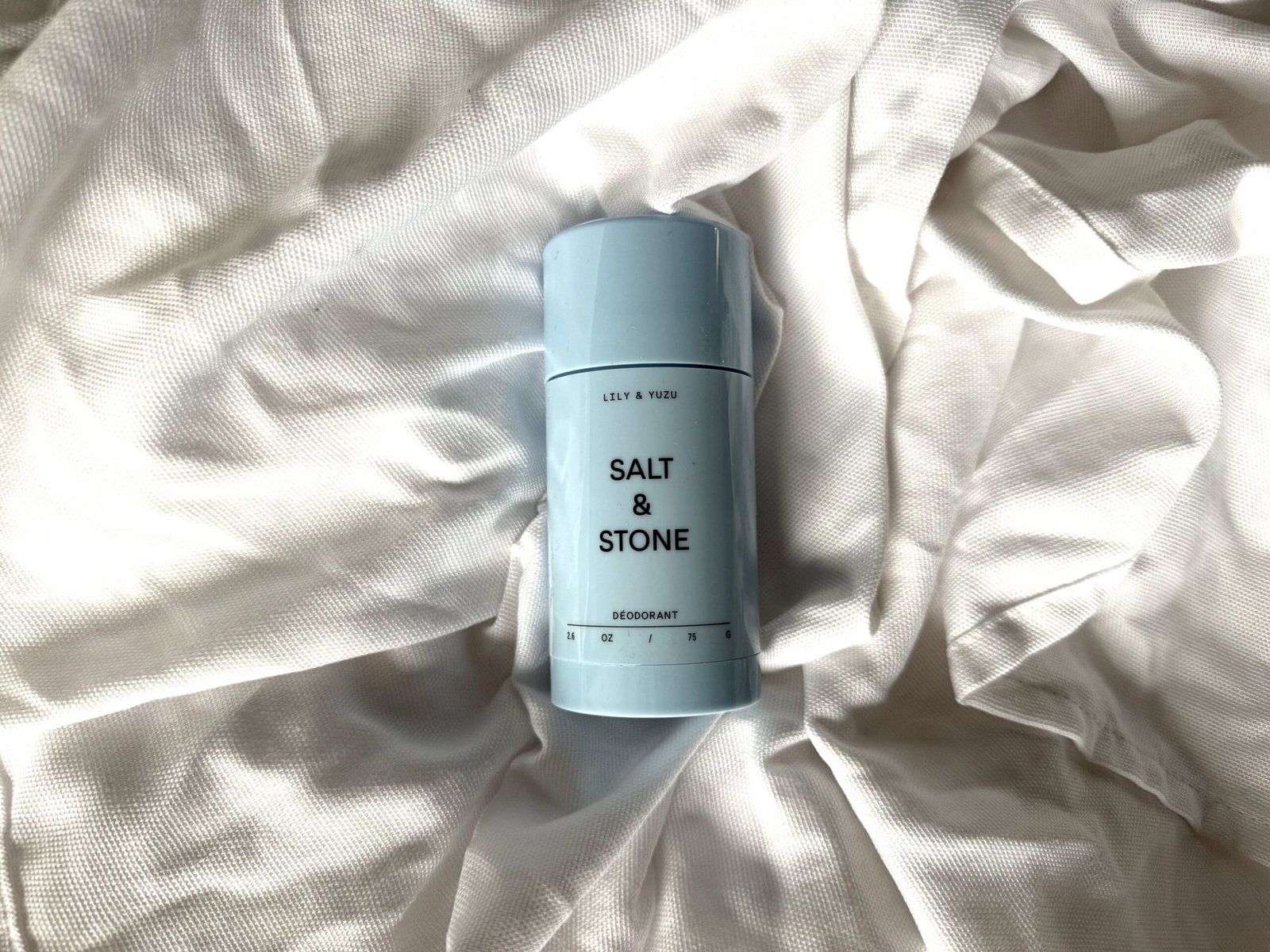 Salt & Stone Lily & Yuzu natural deodorant in pale blue packaging resting on sunlit, textured white fabric.