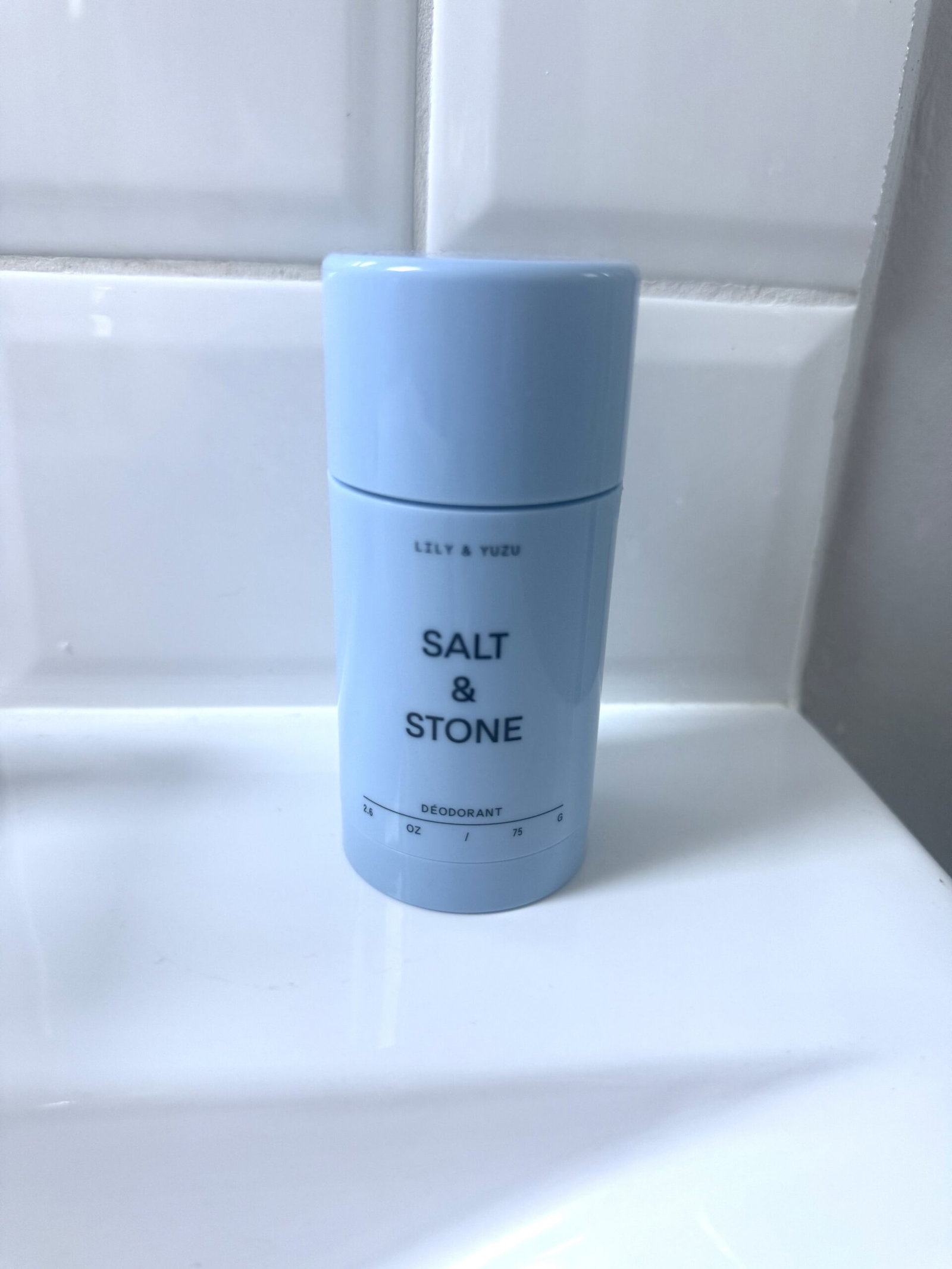 Salt & Stone Lily & Yuzu deodorant standing on a white bathroom shelf against glossy white tiles.