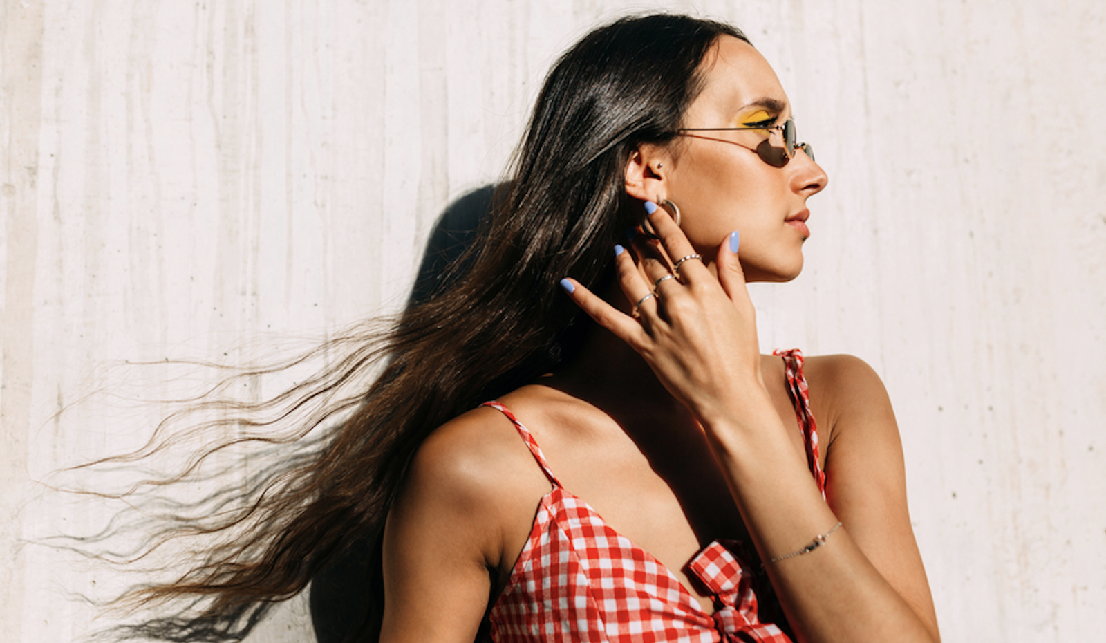 Woman in a red gingham dress and sunglasses standing in bright sunlight, with her long dark hair blowing in the wind.