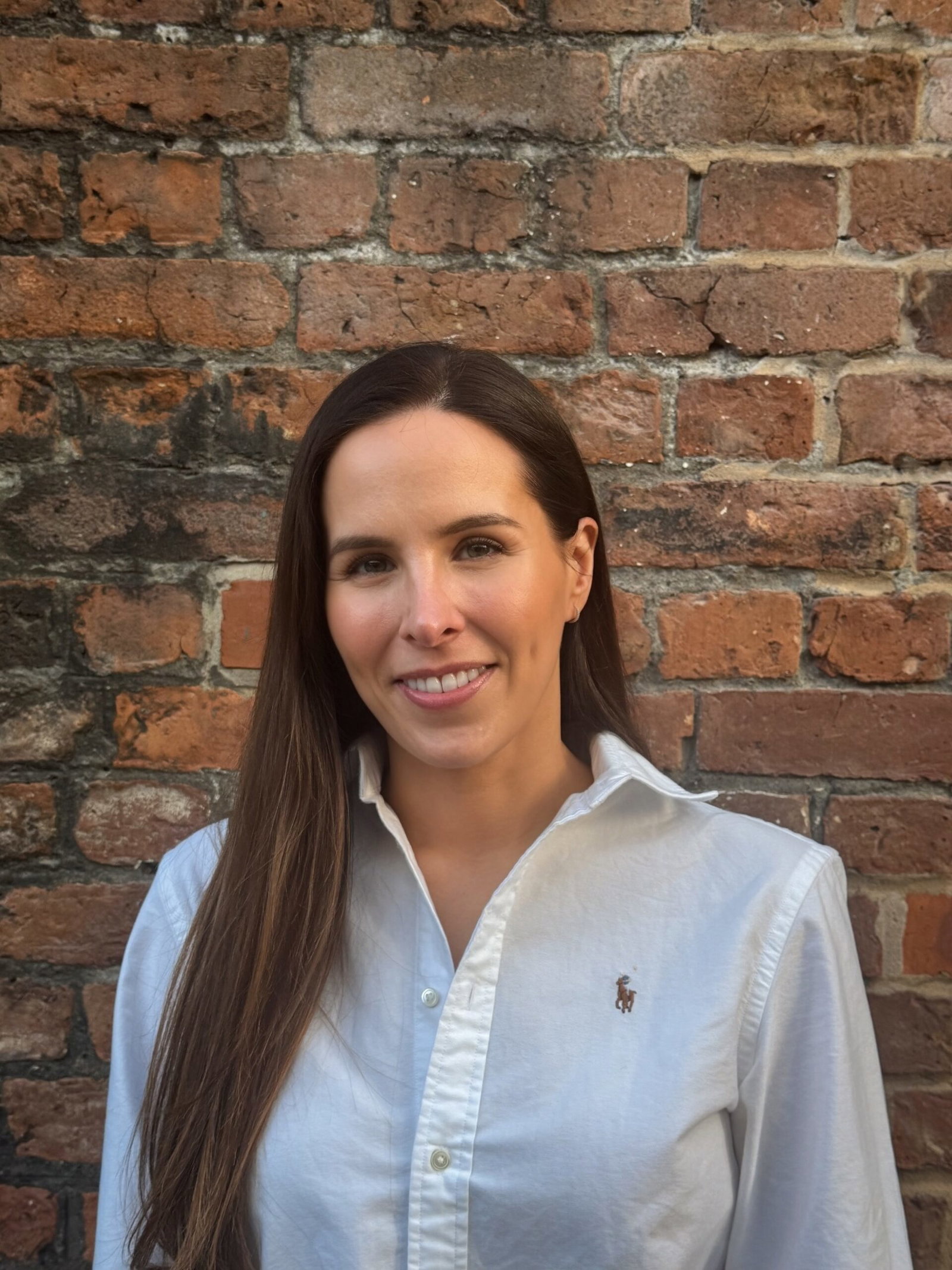 Suzanne Cooper, founder of GLOWWA hair supplements, wearing a white shirt and smiling in front of a red brick wall.