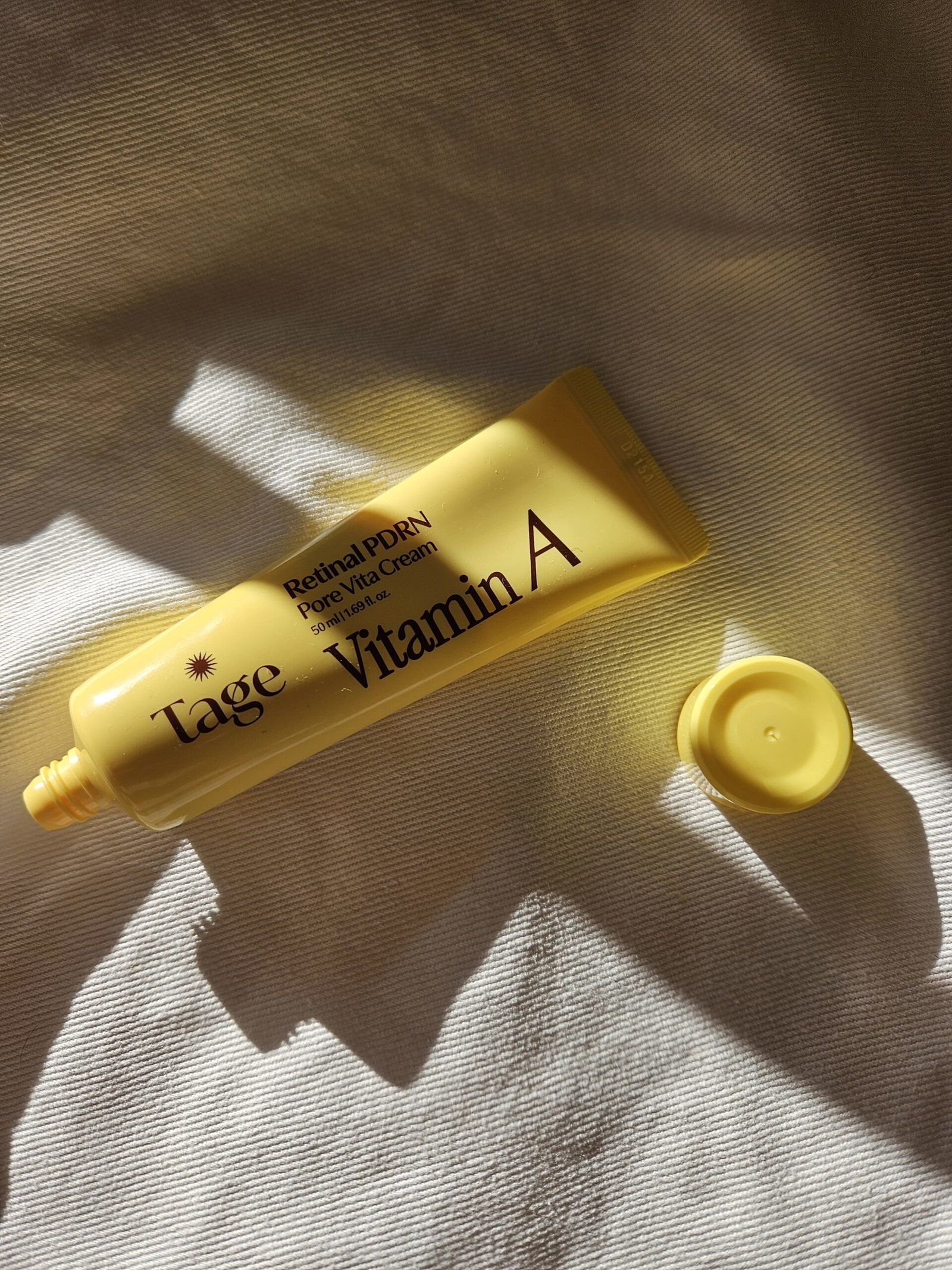 Tage Vita A Retinal PDRN Pore Booster Cream in its yellow tube with the cap removed, photographed on cream fabric in sunlight.