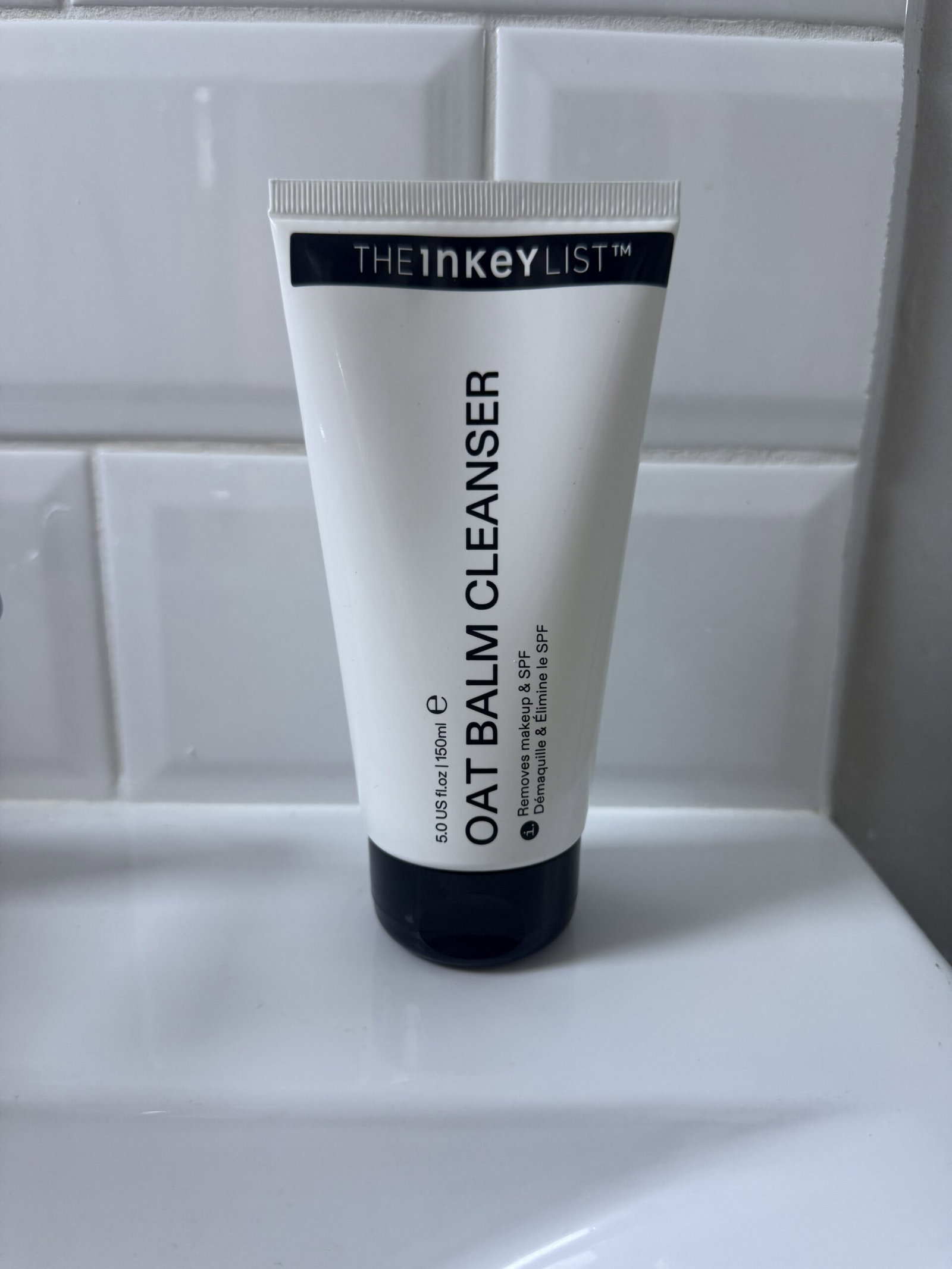 The INKEY List Oat Balm Cleanser in its white and black 150ml tube, photographed against white bathroom tiles.
