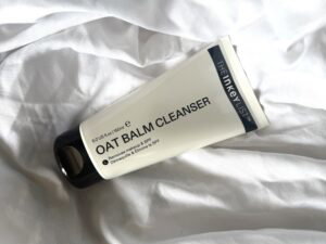 The INKEY List Oat Balm Cleanser in its white tube resting on softly rumpled white bedding, showing the reformulated product packaging.