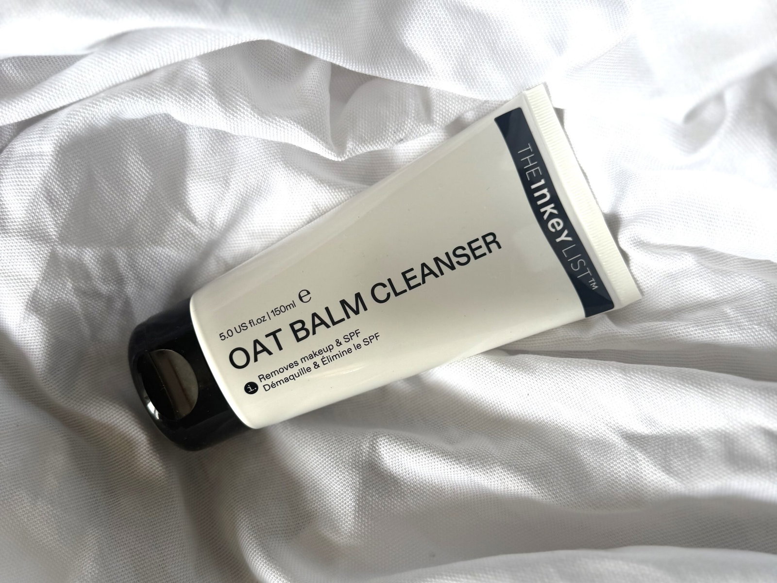 The INKEY List Oat Balm Cleanser in its white tube resting on softly rumpled white bedding, showing the reformulated product packaging.