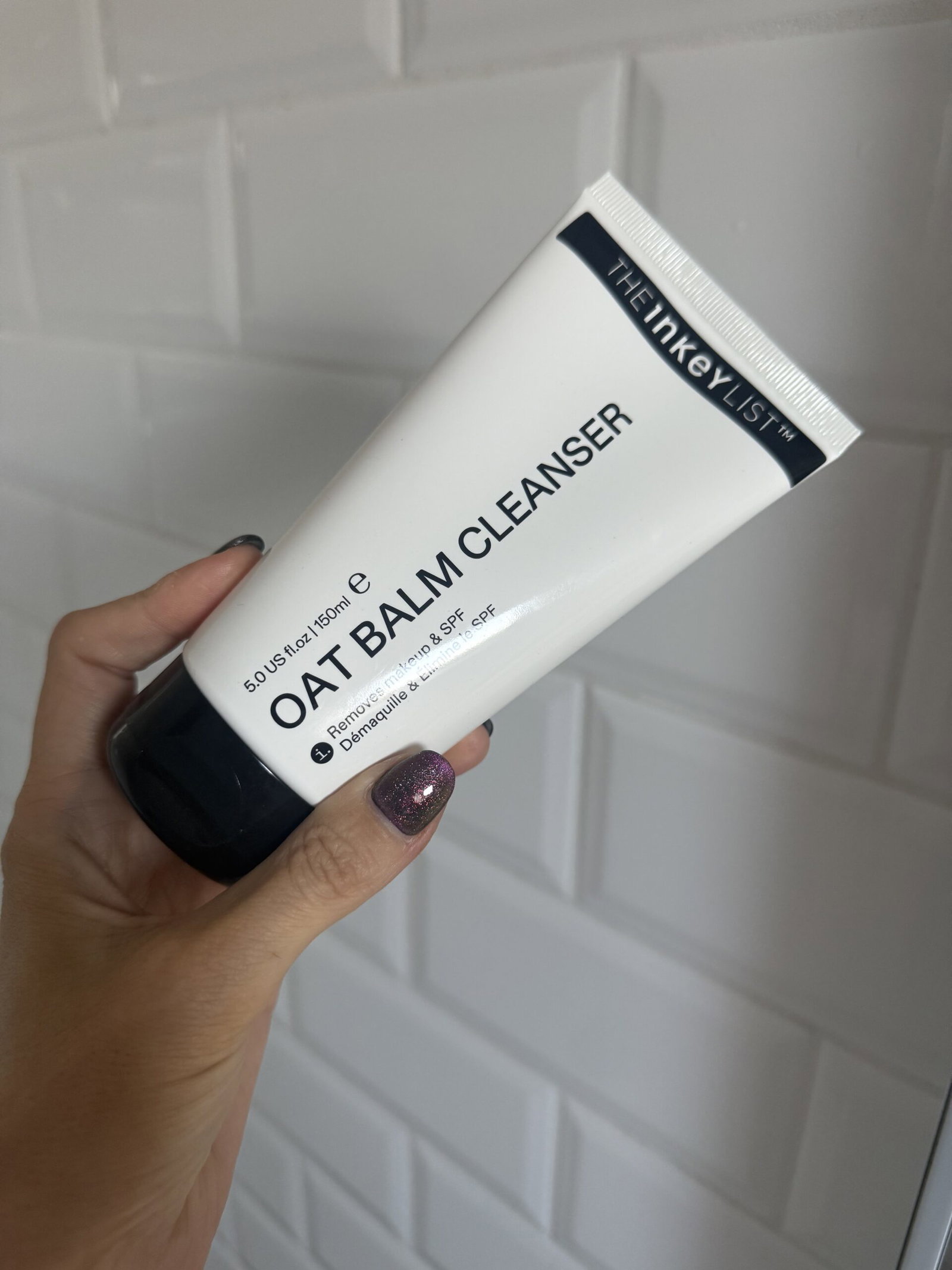 A hand holding the reformulated The INKEY List Oat Balm Cleanser tube against a white tiled bathroom wall.